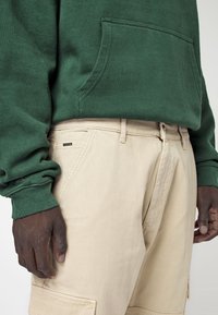 Sudadera con capucha de algodón verde oscuro con bolsillo frontal, combinada con pantalones beige que tienen un bolsillo lateral y una pequeña etiqueta de marca cerca de la cintura.