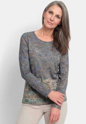 Frau mit langen grauen Haaren, die ein gemustertes Langarmshirt und beige Hosen trägt, steht vor einem schlichten hellen Hintergrund.
