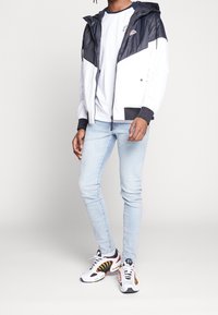 Chaqueta con bloques de color en blanco y azul marino, con capucha, cierre de cremallera y detalles en negro. Combinada con vaqueros azul claro y zapatillas multicolor.