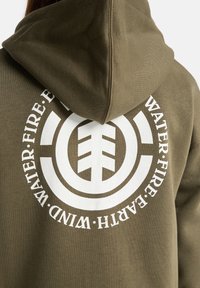 Olivově zelená mikina s velkým bílým kruhovým grafickým motivem na zádech, který obsahuje symboly vody, ohně, země a větru v tučném písmu.