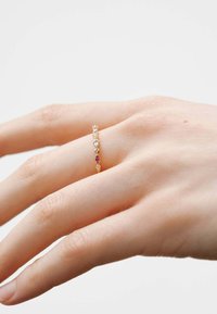 Anillo de oro con múltiples pequeñas piedras preciosas de colores engastadas en una banda delgada, llevado en una mano contra un fondo claro.
