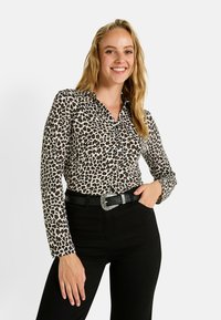 Chemise à imprimé léopard avec des manches longues, en tissu lisse, dotée d'un col et d'une poche avant, associée à un pantalon noir et une ceinture.