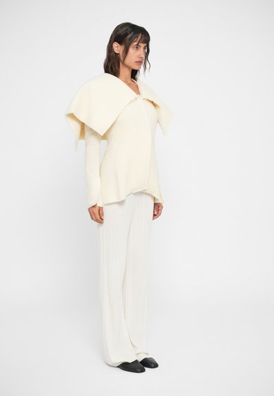 Top de malha creme com colarinho oversized dobrado, combinado com calças largas de malha branca e sapatos pretos de bico fino.