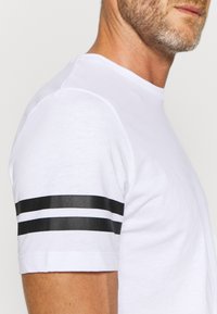 T-shirt bianco in cotone, con scollo rotondo e strisce nere sulle maniche. Tessuto morbido, design semplice e forma aderente.