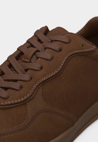 Καφέ σουέντ αθλητικό παπούτσι με ταιριαστά κορδόνια, λεπτομερή ραφή και λεία σόλα σε λευκό φόντο.