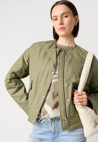 Giacca bomber verde oliva con chiusura a zip, bottoni a pressione e collo a costine. Indossata sopra una camicia a fantasia, abbinata a jeans chiari.