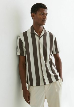 Mand i brun og hvid lodret striped kortærmet skjorte og lyse bukser læner sig op ad hvid væg, kigger til siden med hænderne i lommerne.