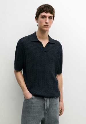 Jeune homme portant un polo en maille à manches courtes bleu marine foncé et un jean gris délavé, debout contre un mur blanc uni.