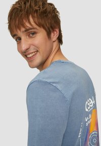 Langsleeve, lichtblauw T-shirt van zacht katoen heeft een grafische print op de achterkant met oranje, gele en witte accenten.