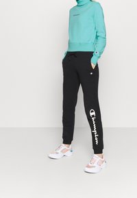 Felpa alta di colore verde acqua con logo ricamato, abbinata a pantaloni jogging neri con stampa del logo bianca. Indossata con scarpe da ginnastica multicolori.