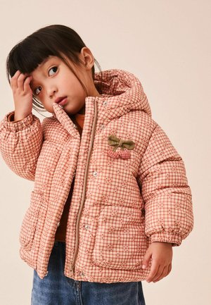 Young child wearing a pink plaid puffer jacket with cherries applique, touching forehead, standing against a plain beige background.