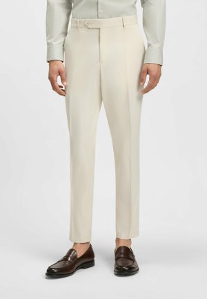 Beige trousers made of smooth fabric, featuring a tailored fit, flat front, and a waistband with a button closure. Paired with brown loafers.