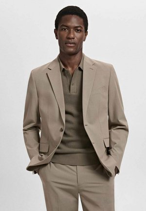 Traje beige de corte ajustado, con un blazer de botón único y un polo texturizado en verde oliva. Tela suave, detalles mínimos.