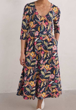 Mujer con un vestido azul marino con estampado floral en rosa, amarillo y verde, mangas tres cuartos, bolsillos laterales y zapatillas blancas.