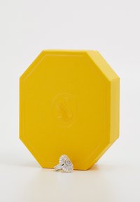 Cutie octagonal galbenă cu o suprafață texturată și logo-ul unui labirint embosat, având o bandă argintie cu un design detaliat care imită scoici.