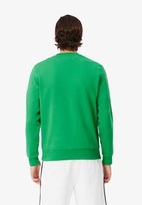 Grøn crewneck sweatshirt med lange ærmer og ribbet kant, lavet af et blødt materiale, båret med hvide bukser med sorte sidestriber.
