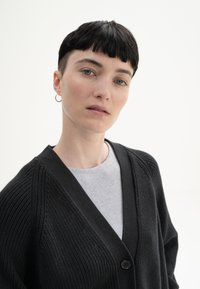 Schwarze Strickjacke mit V-Ausschnitt, Knopfverschluss und strukturiertem Muster, über einem hellgrauen Hemd getragen. Glattes Material.