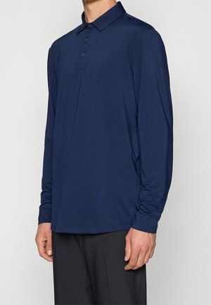 Mann trägt ein marineblaues langärmliges Polohemd und schwarze Hose, steht vor einem schlichten hellen Hintergrund.