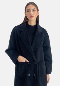 Abrigo de mezcla de lana negro con cuello con muescas, parte delantera de doble botonadura y dos bolsillos laterales. Presenta una superficie suave y texturizada.