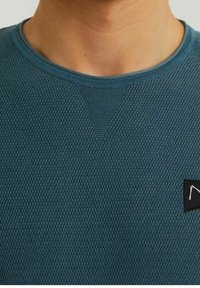 Close-up of a person’s neck and upper chest wearing a textured blue crewneck sweater with a small black logo patch on the right side.