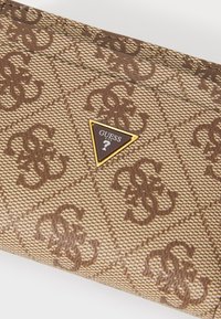 Cartera marrón con patrón, hecha de material sintético texturizado, con un acento triangular dorado que lleva el logo "GUESS".