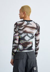 Long-sleeve mesh top featuring a wavy black and white pattern with iridescent accents and a black neckline; logo visible on the back.