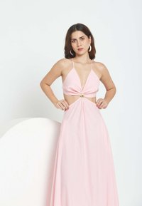 Mujer con cabello oscuro de longitud media, vestida con un vestido sin mangas color rosa claro con recortes y un detalle de anillo dorado en la cintura, de pie con las manos en las caderas.
