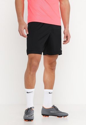 Person iført sorte Nike-shorts, hvide Nike-strømper, grå fodboldstøvler med orange knopper og en lyserød sportsbluse, stående foran en hvid baggrund.
