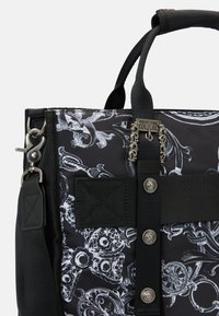 Sac en tissu noir avec des motifs ornementaux blancs, des accessoires en argent, des sangles noires contrastantes et un détail de logo avec des éléments décoratifs.