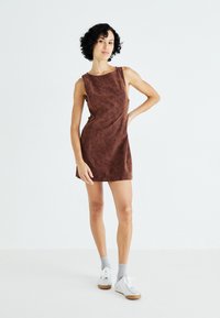 Bruine mouwloze jurk met een zachte, gestructureerde afwerking. Beschikt over een iets losse pasvorm en een zijsplit, gecombineerd met lichtgrijze sportsokken en sneakers.
