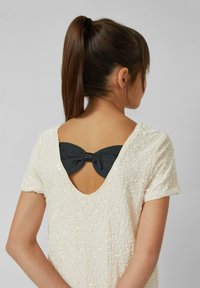 Kremowy top pokryty małymi cekinami, z głębokim dekoltem V z tyłu ozdobionym czarną kokardą. Krótkie rękawy i luźny fason.