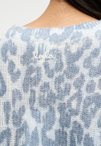 Κοντινή εικόνα του πίσω μέρους του λαιμού ενός χνουδωτού πλεκτού πουλόβερ με μπλε και λευκή λεοπάρδαλη και ένα μικρό κεντημένο λογότυπο "lala Berlin".