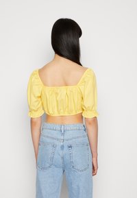 Blusa amarela cropped com mangas bufantes e punhos elásticos, apresentando um decote quadrado nas costas. Combinada com jeans de cintura alta em azul claro.