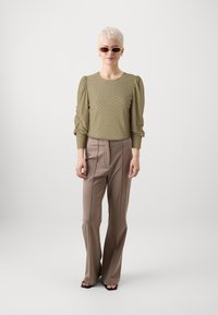 Olive green knit top with intricate patterns and puff sleeves, paired with tan high-waisted trousers. Model wearing brown sunglasses and black heels.