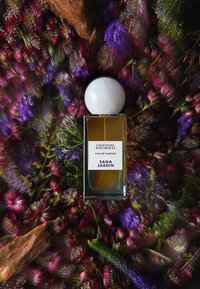 Bottiglia di eau de parfum con una forma rettangolare trasparente, liquido dorato e tappo sferico bianco, circondata da fiori vibranti di colore viola e rosa.