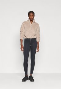 Pull en polaire beige à demi-zippé avec un ourlet à cordon, associé à des leggings noirs et des chaussures de sport noires. Textures lisses et douces.