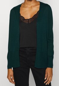 Cárdigan verde oscuro con frente abierto y puños acanalados. Usado sobre una camiseta de encaje negra, que presenta una textura de punto suave y sin patrones visibles.