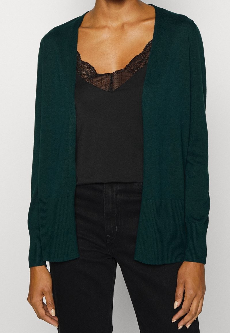 Cárdigan verde oscuro con frente abierto y puños acanalados. Usado sobre una camiseta de encaje negra, que presenta una textura de punto suave y sin patrones visibles.