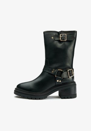 Black leather ankle boot with a rounded toe, matte finish, thick sole, and gold-tone buckles. Features a strap across the midfoot.