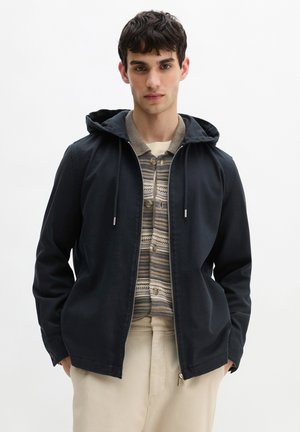 Jonge man die een zwart capuchonjack draagt over een beige vest met patroon en lichtgekleurde broek, staand met handen in de zakken.