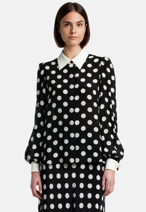Blusa nera con grandi pois bianchi, caratterizzata da un colletto bianco, chiusura a bottoni e maniche a sbuffo. Tessuto liscio con una finitura lucida.
