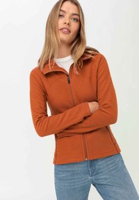 Orangene Fleecejacke mit Reißverschluss, hohem Kragen und langen Ärmeln. Hat ein figurbetontes Design und eine glatte Textur. Wird über einem weißen Hemd getragen.