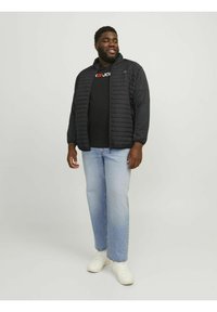 Sort quilted jakke med lange ærmer, båret over en sort t-shirt, parret med lyseblå jeans og hvide sneakers, stående mod en lys baggrund.