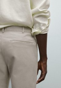 Beige cotton trousers with a sleek design, featuring a single back pocket and a smooth texture, paired with a light-colored linen shirt.