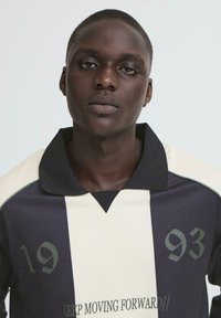 Mørkeblå og creme-stribet rugbytr øje med sort krave, der har tallene 19 og 93 samt teksten "FREMSKRIDT UDAD".