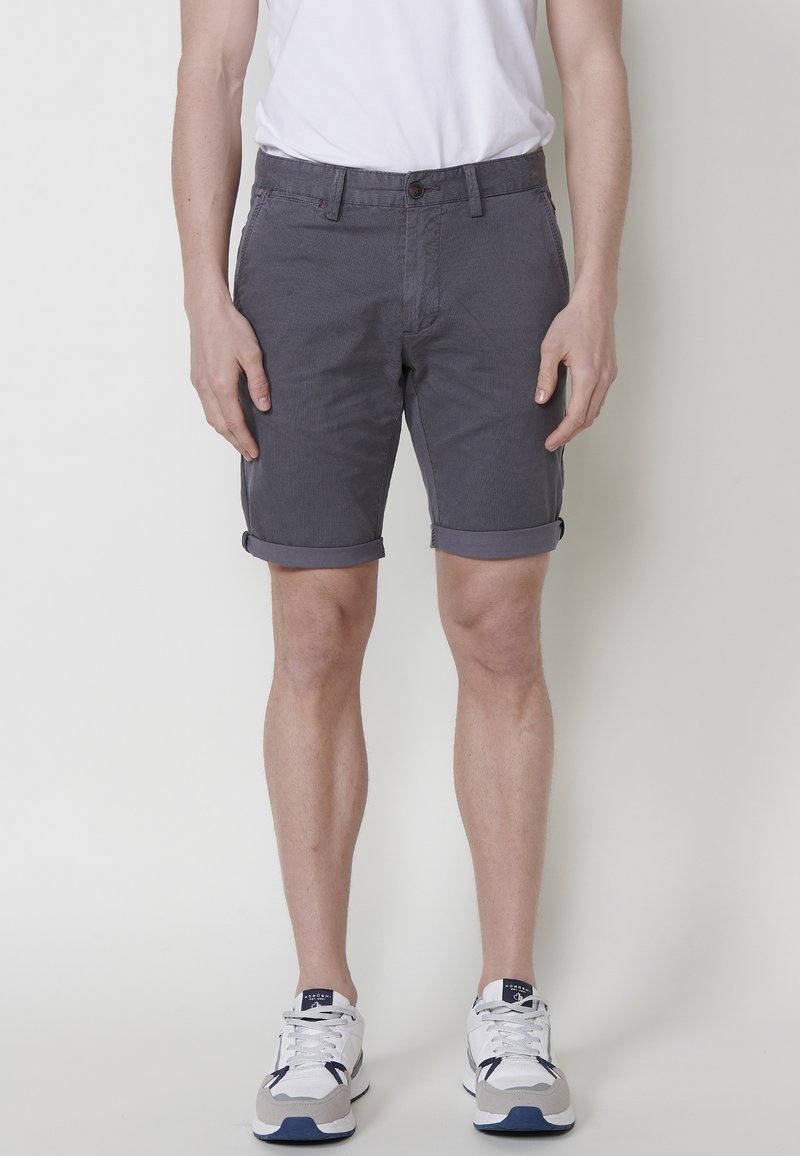 Férfi, aki felhajtott szürke sortot, fehér pólót és fehér sportcipőt visel, egy sima világos háttér előtt áll.