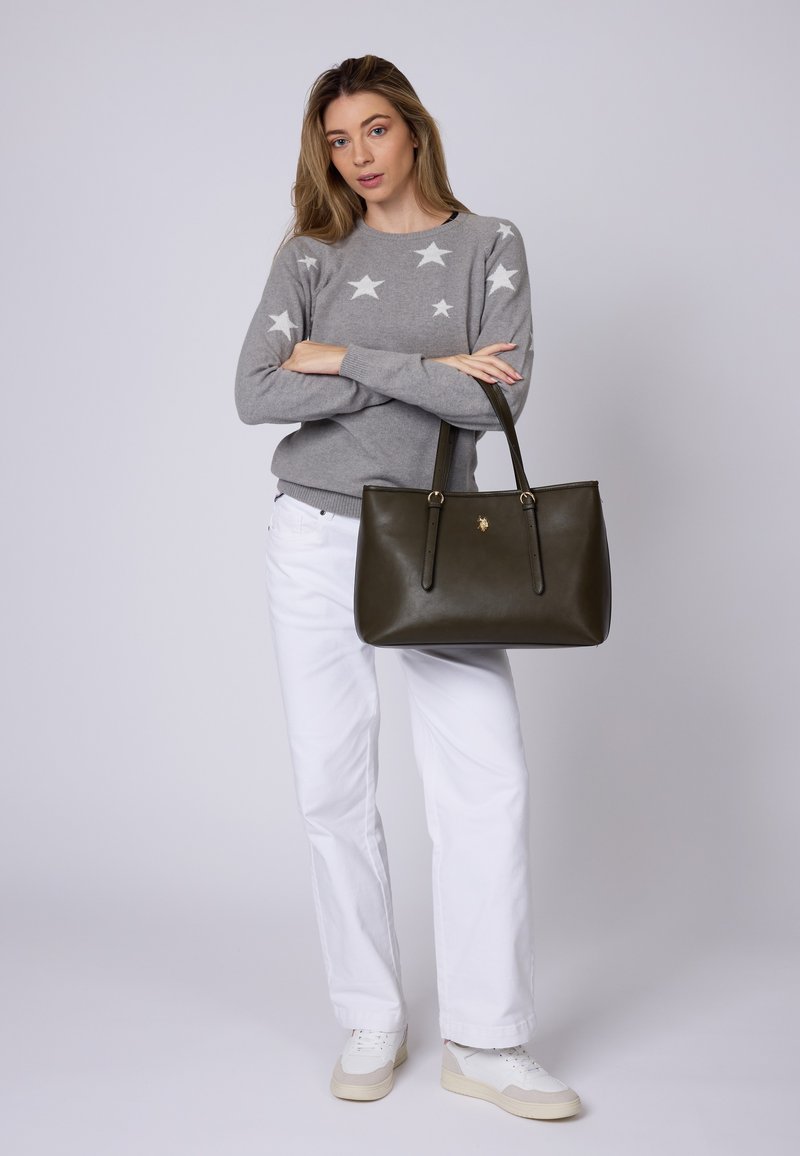 Borsa a tracolla in pelle verde oliva con doppi manici e un emblema dorato. Il modello indossa un maglione grigio con motivi a stelle bianche e pantaloni bianchi.