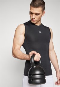 Black sleeveless athletic top with a light logo, held by a person grasping a black kettlebell. Smooth, fitted material; muscular structure visible.