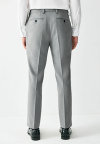 Pantalones a medida grises con un tejido texturizado, pinzas frontales y bolsillos traseros con cierres de botón. Combinados con zapatos negros.