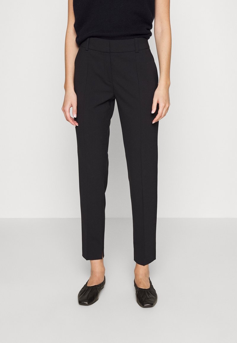 Femme portant un pantalon noir slim longueur cheville, un haut noir sans manches et des chaussures plates noires, debout devant un fond uni.